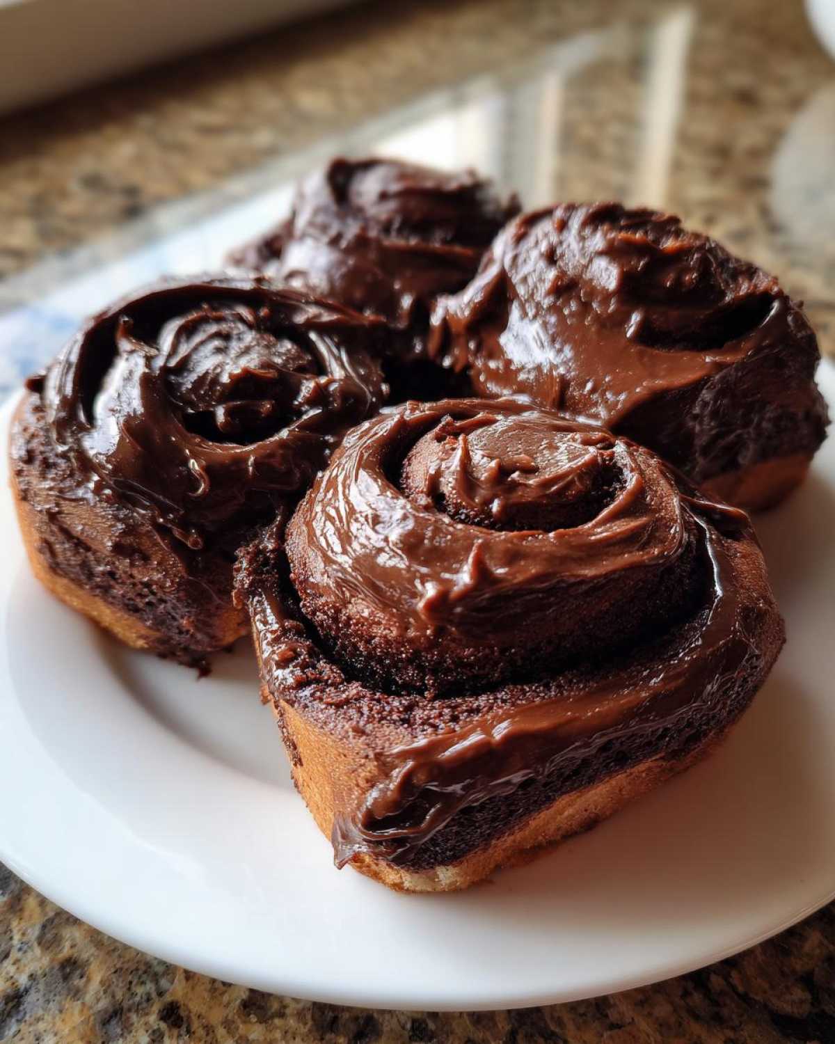 Four rich chocolate cinnamon rolls heavily frosted with thick, glossy chocolate icing on a white plate.