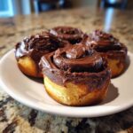 Four freshly baked chocolate cinnamon rolls generously topped with thick, glossy chocolate frosting on a white plate.