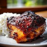 Close-up of a perfectly cooked piece of harissa chicken with a dark, caramelized glaze next to a serving of fluffy white rice.