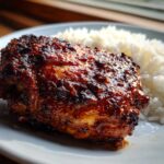 Close-up of a piece of richly glazed, dark red harissa chicken next to a serving of fluffy white rice.