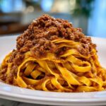 Close-up of rich bolognese sauce piled high on fresh, yellow tagliatelle pasta on a white plate.