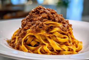 Close-up of rich bolognese sauce piled high on fresh, yellow tagliatelle pasta on a white plate.
