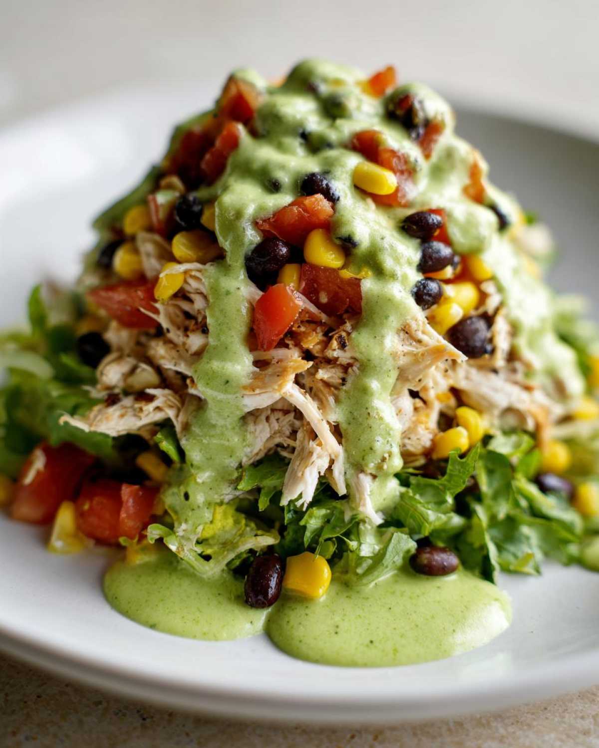 A tall serving of southwest chicken salad featuring shredded chicken, lettuce, black beans, corn, tomatoes, and a generous drizzle of green avocado dressing.