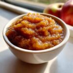 A close-up, appetizing shot of thick, spiced applesauce in a white bowl, with fresh apples blurred in the background.