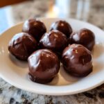A close-up of six rich, dark chocolate covered Oreo balls arranged on a white plate.