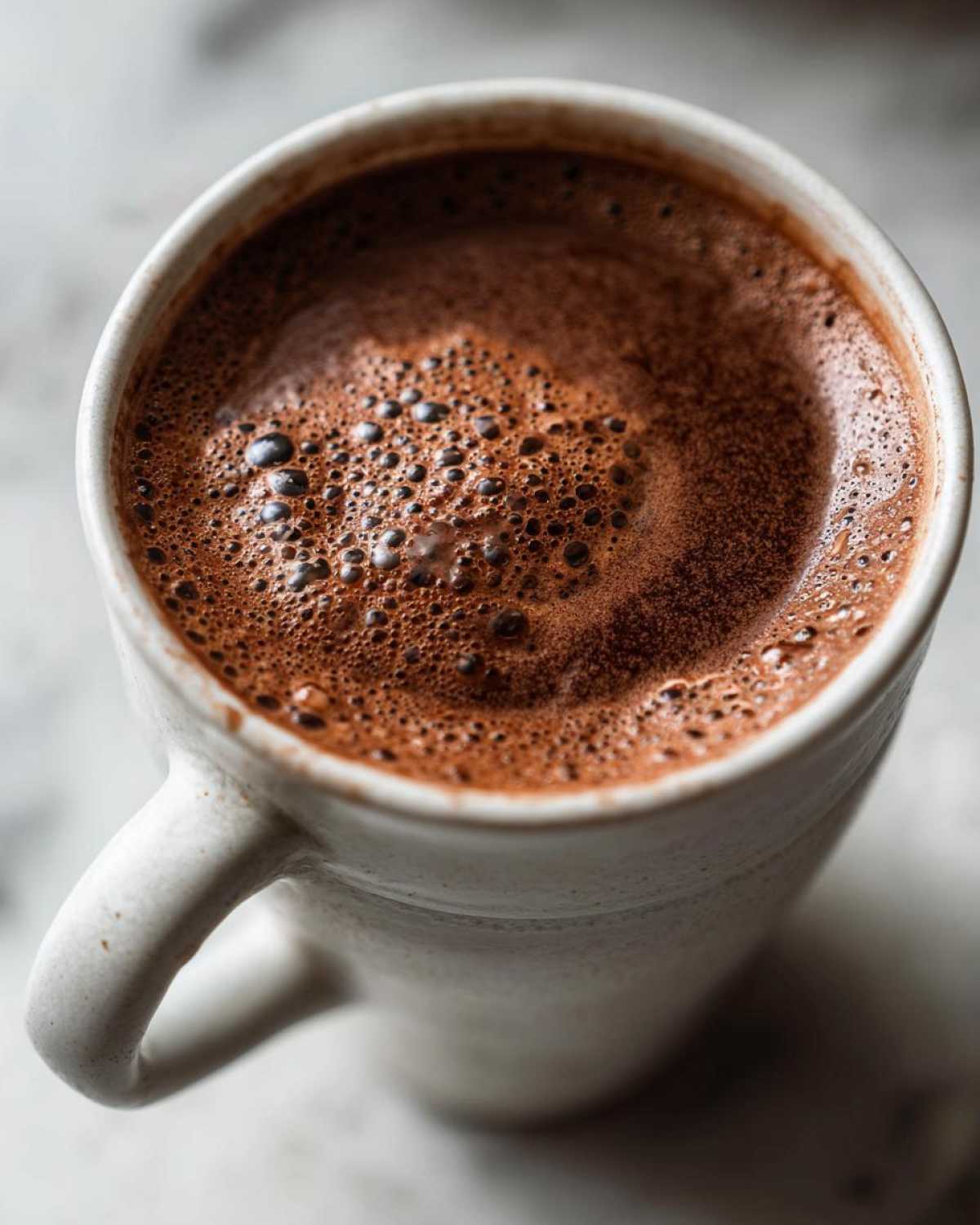 Close-up overhead view of rich, foamy homemade hot cocoa in a textured white mug.