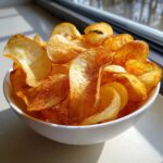 A close-up of crispy, seasoned potato chips piled high in a white bowl near a window.
