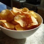 A white bowl overflowing with golden, seasoned homemade potato chips, catching bright sunlight.