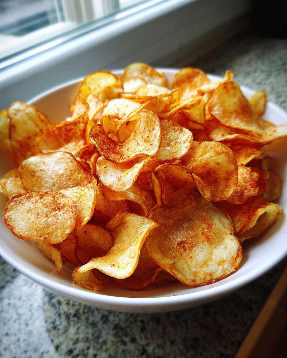 A white bowl filled with crispy, golden, seasoned homemade potato chips, sitting near a window.