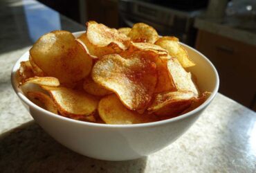 A white bowl overflowing with golden, seasoned homemade potato chips, catching bright sunlight.