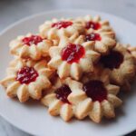 A pile of freshly baked, star-shaped Italian butter cookies filled with bright red jam.