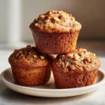 Three golden brown banana nut muffins stacked on a white plate, topped generously with chopped walnuts.
