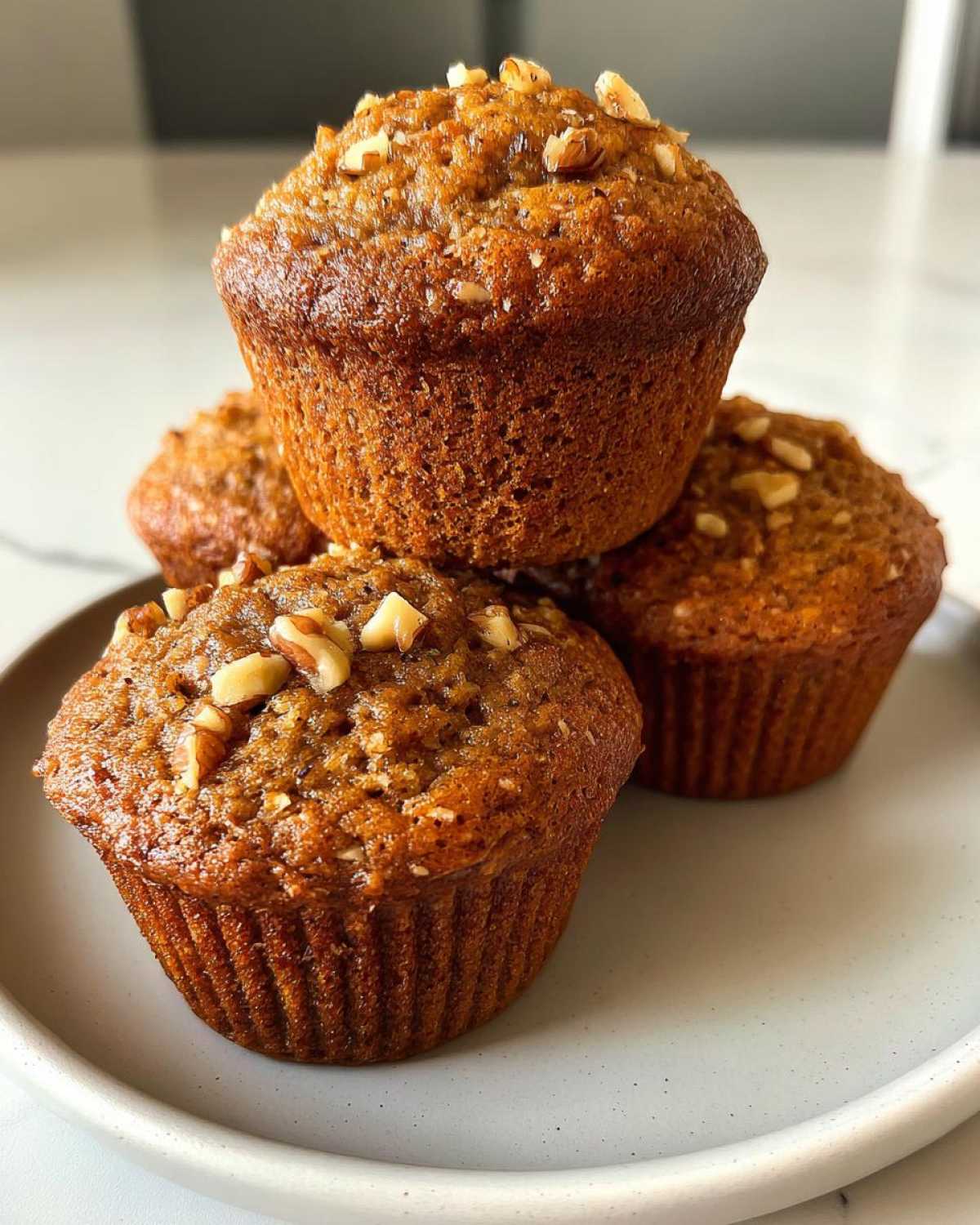 A stack of four golden brown banana nut muffins topped with chopped walnuts sitting on a light gray plate.