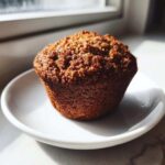 A single, dark brown, freshly baked bran muffin sitting on a small white saucer near a sunlit window.
