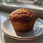 A single, perfectly baked, moist bran muffin sitting on a small ceramic plate, highlighted by bright sunlight.