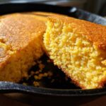 Close-up of a slice of golden buttermilk cornbread with a crumbly texture, sitting in a black cast iron skillet.