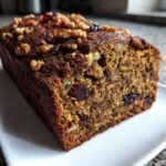 A close-up of a slice of moist date nut bread loaf topped with walnuts and visible dried fruit.