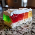 A cross-section slice of a colorful jello poke cake showing layers of white cake, vibrant green, red, orange, and yellow gelatin, topped with whipped cream.