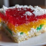 A close-up of a moist slice of jello poke cake featuring vibrant rainbow layers of gelatin over a white cake base, topped with whipped cream.