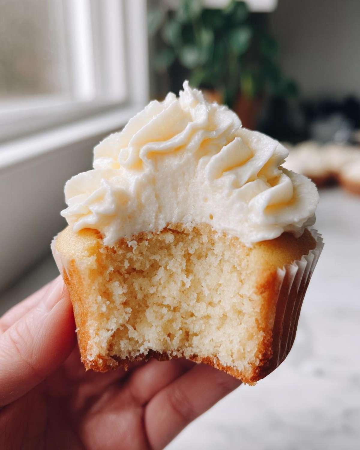 Close-up of a hand holding a vanilla cupcake with a bite taken out, showing the moist crumb and creamy frosting.
