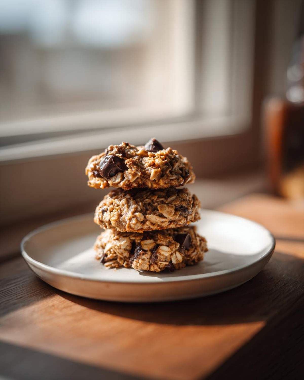 Three stacked oatmeal chocolate chip breakfast cookie on a small plate near a window.