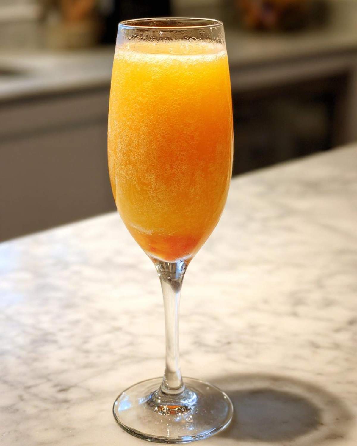 A tall flute glass filled with a vibrant orange, bubbly bellini recipe mixture resting on a marble countertop.