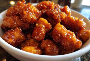 Close-up of crispy pieces of orange chicken recipe coated in a thick, shiny orange glaze and sprinkled with sesame seeds.