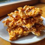 A stack of golden, glossy pieces of homemade peanut brittle recipe resting on a white plate near a window.