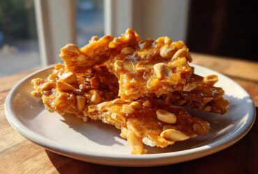 Pieces of golden, glossy peanut brittle recipe stacked on a small white plate, catching the sunlight.