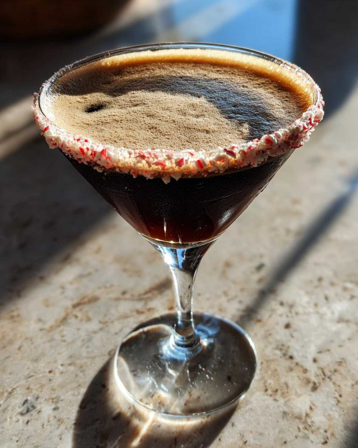 A rich, dark peppermint espresso martini in a coupe glass with a crushed candy cane rim, sitting on a marble counter.