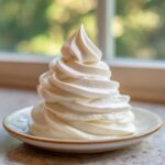 A tall, perfectly piped swirl of white, fluffy cream cheese frosting served on a small saucer.