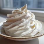 A beautiful, tall swirl of fluffy white cream cheese frosting piped onto a small, rustic saucer.