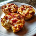 Three golden-brown pizza muffins on a white plate, one has a bite taken out, showing the fluffy interior.