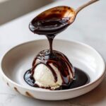 A spoonful of thick, glossy chocolate syrup is being poured over a scoop of vanilla ice cream in a white bowl.