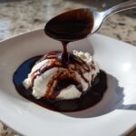 A spoonful of rich chocolate syrup is drizzled over a scoop of vanilla ice cream in a white bowl.