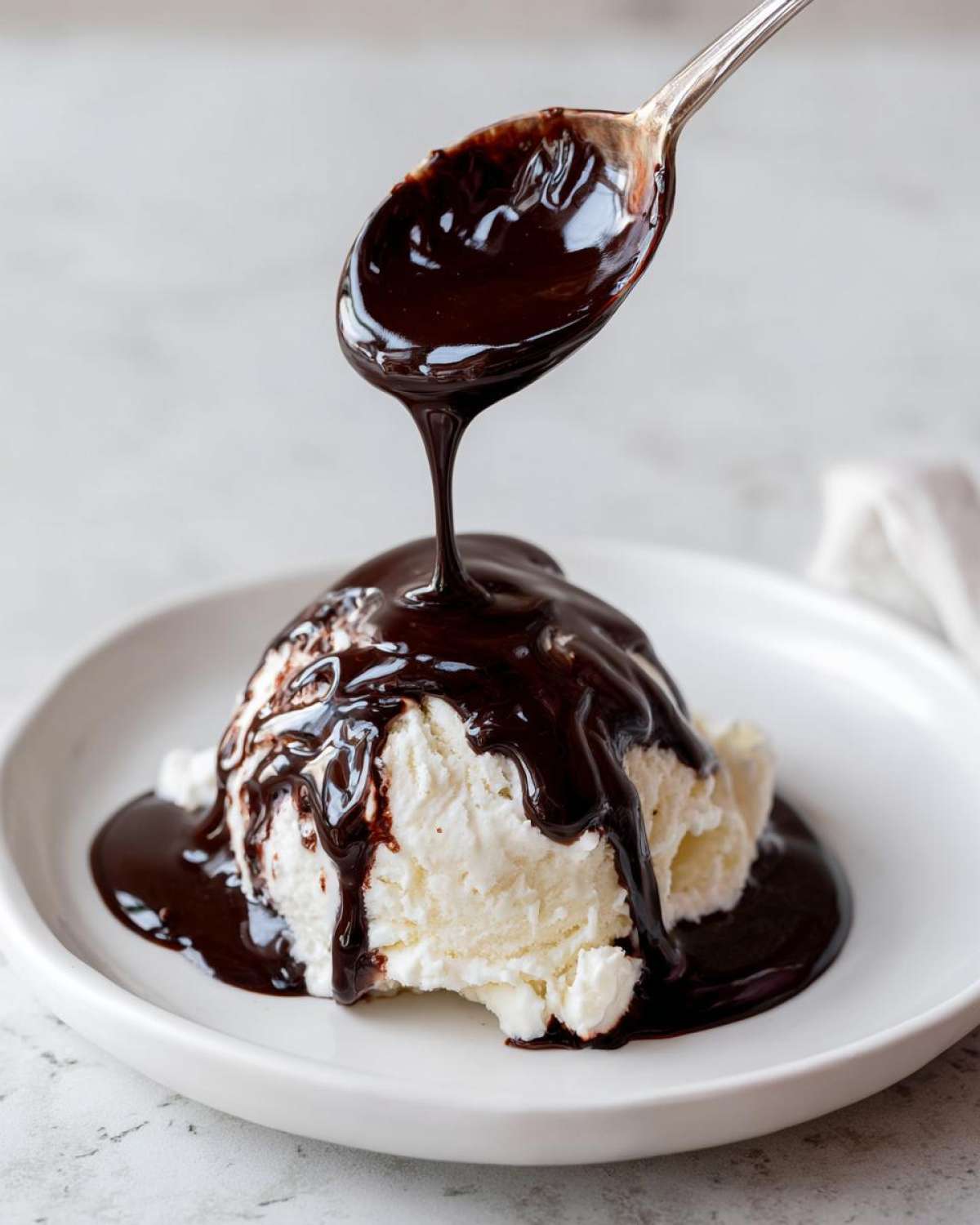 A spoonful of thick, glossy chocolate syrup is being poured over a scoop of vanilla ice cream.