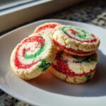 A stack of colorful red and green pinwheel cookies dusted with sugar, sitting on a white plate near a window.