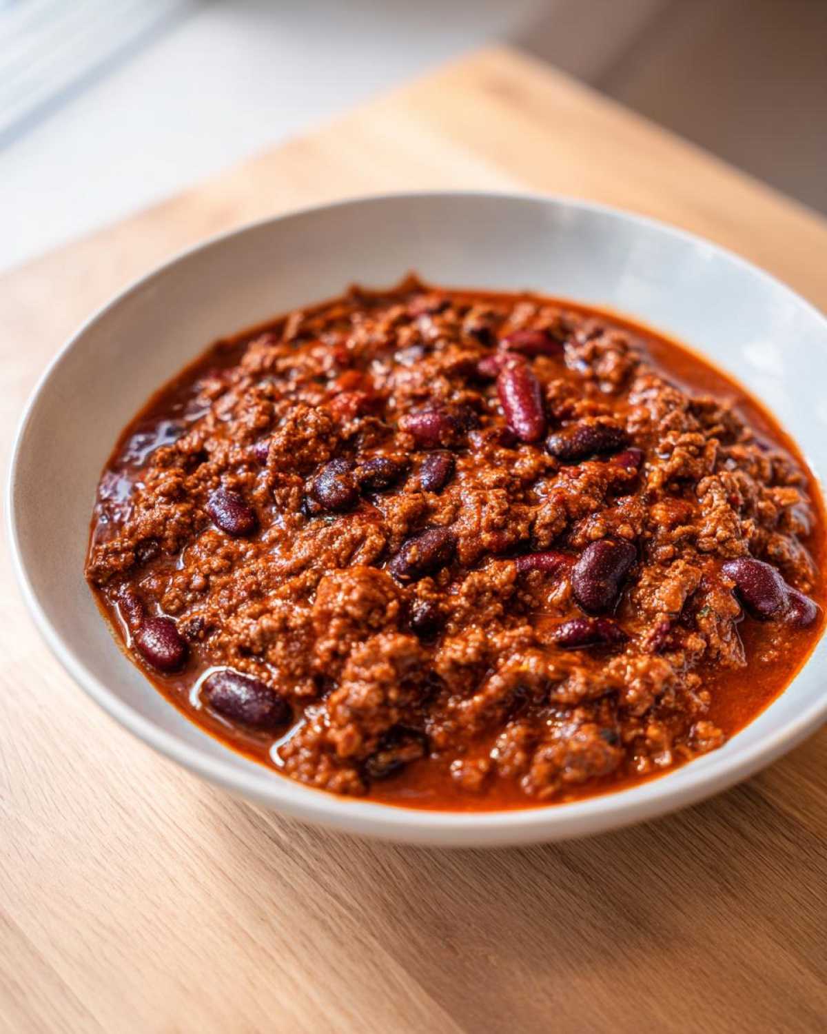 A close-up view of a white bowl filled with rich, thick crockpot chili featuring ground meat and kidney beans.