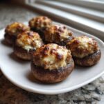 Six golden-brown sausage stuffed mushrooms with melted, browned cheese topping, served on a white plate.