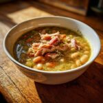 A close-up of a savory 1 pot bean soup topped with shredded smoked ham, served in a white bowl on a rustic wooden surface.