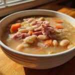 A close-up of a white bowl filled with savory ham and white bean soup, featuring carrots and celery.