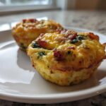Two savory egg bites, featuring bacon and spinach baked in a muffin tin, sitting on a white plate. Perfect for a quick egg breakfast.