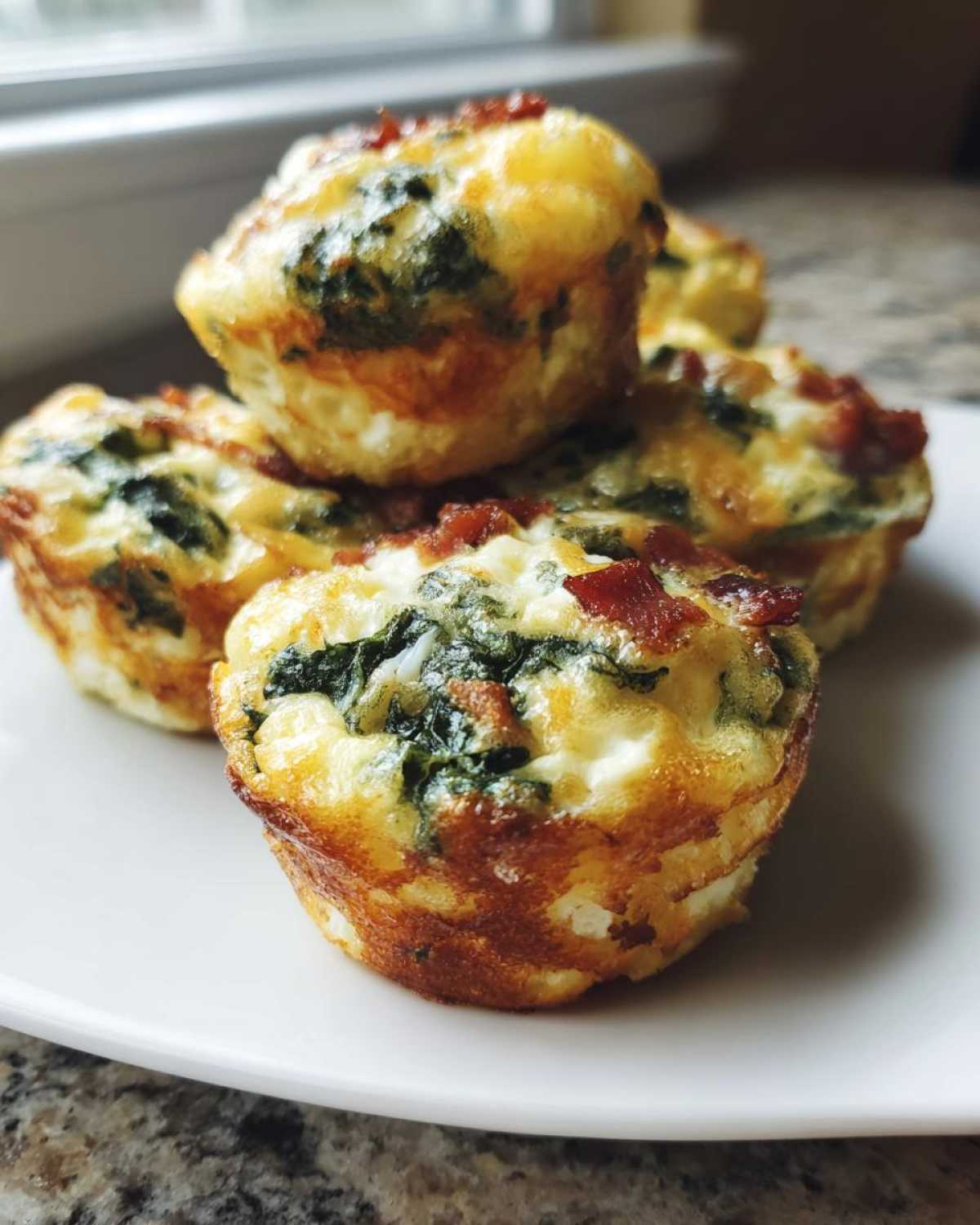 A stack of five golden-brown baked egg bites mixed with spinach and bacon pieces, served on a white plate.
