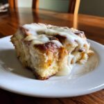 A close-up of a square serving of gooey Cinnamon roll casserole topped with thick white icing on a white plate.