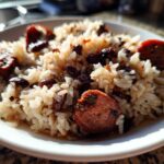 Close-up of a white bowl filled with seasoned white rice, black beans, and slices of smoked sausage, representing black beans and rice.