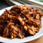 Close-up of shredded crockpot teriyaki chicken coated in a rich, glossy sauce served on a white plate.