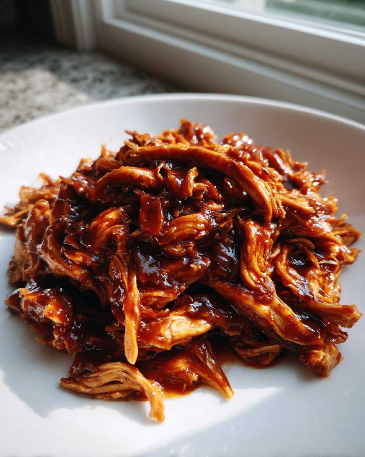 Close-up of shredded crockpot teriyaki chicken coated in a thick, glossy, dark teriyaki sauce, served on a white plate.
