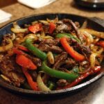 Close-up of sizzling steak fajitas with sliced beef, red peppers, green peppers, and onions in a black cast iron skillet.
