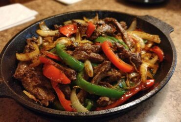 Close-up of sizzling steak fajitas with sliced beef, red peppers, green peppers, and onions in a black cast iron skillet.