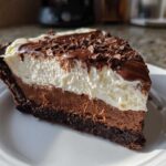 A close-up slice of a 4-layer mud pie showing chocolate crust, mousse, whipped cream, and chocolate shavings.